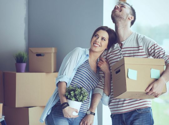 Un couple heureux aménage dans un appartement : ils portent un carton et une plante verte