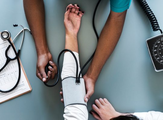 Un soignant prenant la tension d'une personne. Toutes les deux sont assises à un bureau, sur celui ci se trouve des papier et un stéthoscope