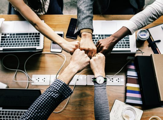 Le travail en équipe : un groupe de personne se tapant les poings en formant une étoile au-dessus d'un bureau sur lequel est posée trois ordinateurs