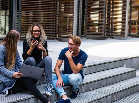 Un groupe de trois personnes assis sur des marches : la personne de gauche a unordinateur sur les genoux, celle du milieu son téléphone à la main. Ils discutent.