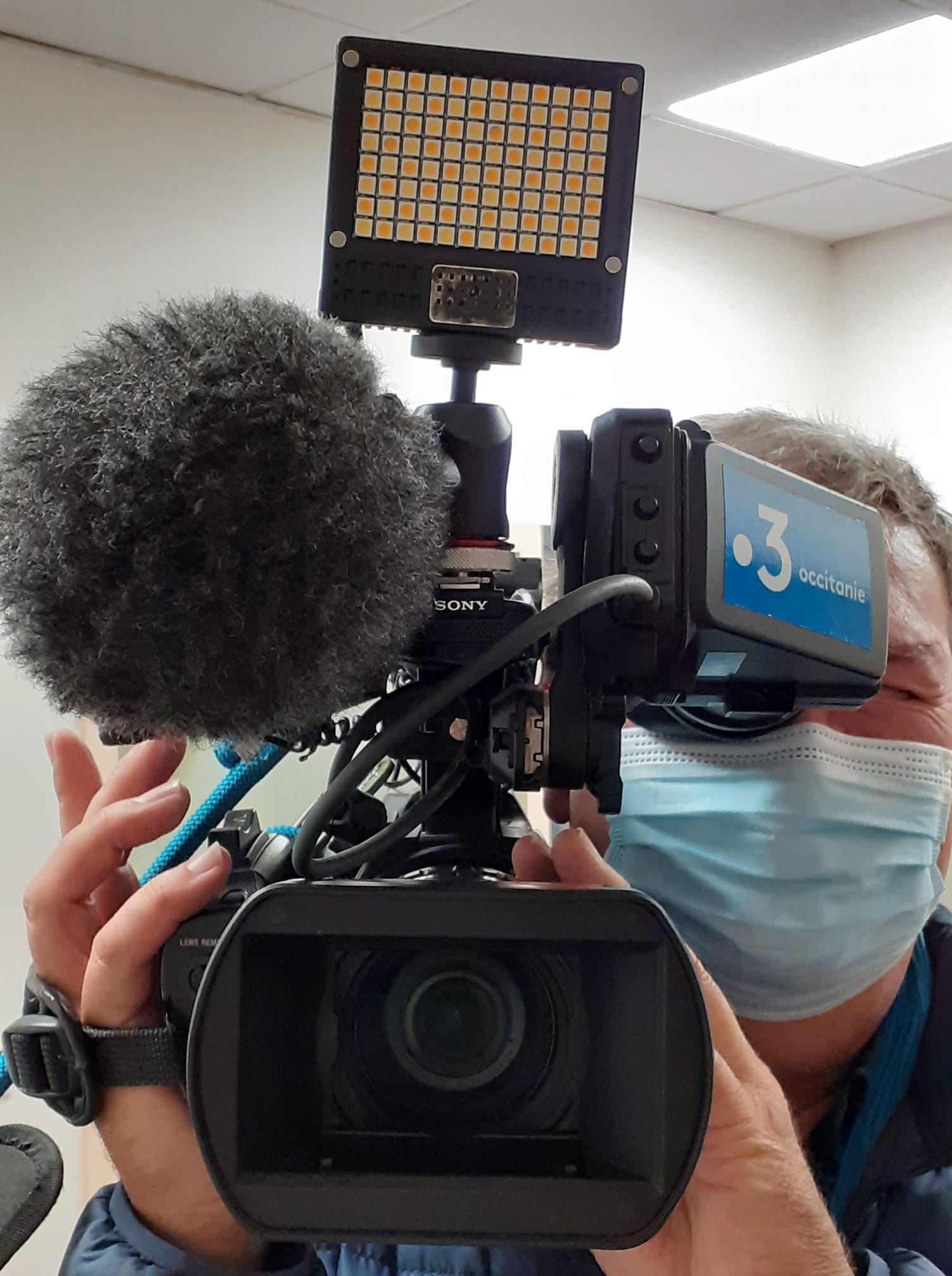 De face une caméra de France 3 Occitane avec un micro, en arrière plan le caméraman